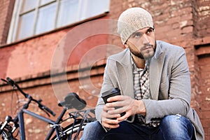 Handsome young man in grey coat and hat sitting on a bench relaxed drinking coffee and thinking near his bicycle