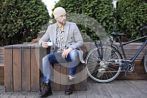 Handsome young man in grey coat and hat sitting on a bench relaxed drinking coffee and thinking near his bicycle