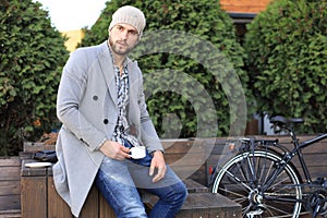 Handsome young man in grey coat and hat sitting on a bench relaxed drinking coffee and thinking near his bicycle