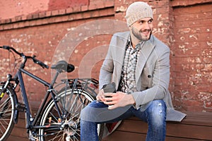 Handsome young man in grey coat and hat sitting on a bench relaxed drinking coffee and thinking near his bicycle