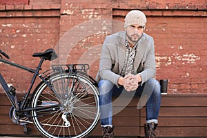 Handsome young man in grey coat and hat sitting on a bench relaxed drinking coffee and thinking near his bicycle