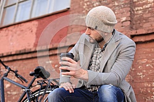 Handsome young man in grey coat and hat sitting on a bench relaxed drinking coffee and thinking near his bicycle