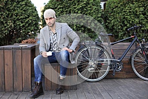 Handsome young man in grey coat and hat sitting on a bench relaxed drinking coffee and thinking near his bicycle