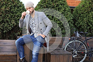 Handsome young man in grey coat and hat sitting on a bench relaxed drinking coffee and thinking near his bicycle