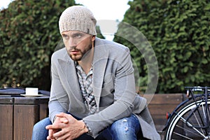 Handsome young man in grey coat and hat sitting on a bench relaxed and thinking near his bicycle outdoors