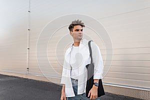 Handsome young man with a hairstyle in a fashionable shirt in blue jeans with a black cloth bag poses in a modern building