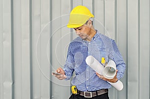 Handsome young man in hardhat holding blueprint and using mobile phone while standing outdoors and against building structure