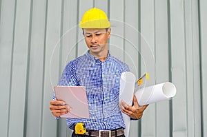 Handsome young man in hardhat holding blueprint and using tablet while standing outdoors and against building structure