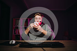 A handsome young man in a headset sits at night on a computer at home with a cup of tea in his hands and looks at the camera with