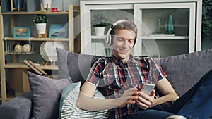 Handsome young man hipster is listening to music through headphones, using smartphone touching screen and laughing