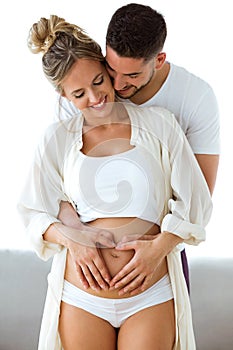 Handsome young man holding belly of his pregnant wife making heart together at home
