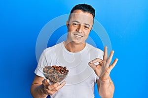 Handsome young man holding bowl of star anise doing ok sign with fingers, smiling friendly gesturing excellent symbol