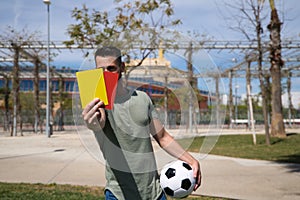 Handsome young man holding a football in his hand while showing red and yellow card to camera as if he were a football referee.