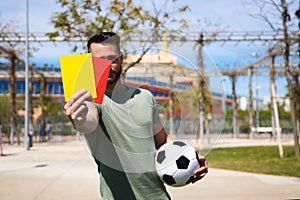 Handsome young man holding a football in his hand while showing red and yellow card to camera as if he were a football referee.
