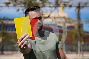 Handsome young man holding a football in his hand while showing red and yellow card to camera as if he were a football referee.