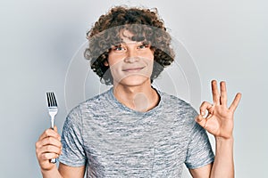 Handsome young man holding one silver fork doing ok sign with fingers, smiling friendly gesturing excellent symbol