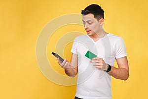 handsome young man holding a phone and a bank card in his hand in a white t-shirt on a yellow background