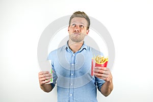 Handsome young man holds soda in papper cup and fries from fast food restaurant looks doubtful on isolated white