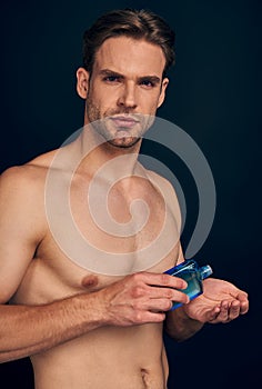 Handsome young man isolated. Portrait of shirtless muscular man is standing on dark blue background and using face aftershave