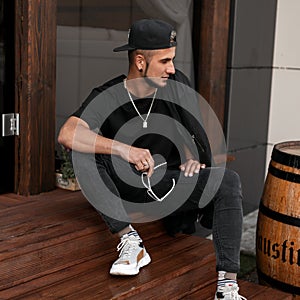 Handsome young man in jeans in a trendy cap in a black T-shirt with a jacket with sunglasses with a beard is sits