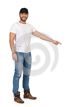 Handsome Young Man In Jeans, White T-shirt And Black Cap Is Looking At Camera And Pointing Down