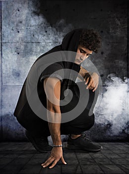 Handsome young man kneeling down in dark hoodie on black background