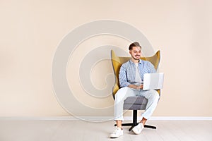 Handsome young man with laptop in armchair near color wall indoors