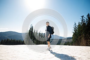 Handsome young man in light clothes stands in the snow in the mountains with a backpack on his back, posing for the camera while