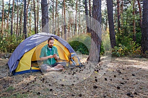 Handsome young man listening to music in headphones while sitting in yellow grey tent, enjoying nature in autumn forest