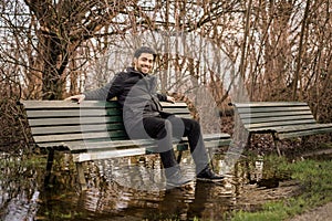 Handsome young man looking at camera while sitting on a flooded park bench in winter