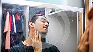 Handsome young man looking at himself at the mirror in wardrobe room while touching his beard