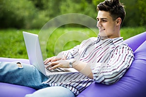 Handsome young man lying on inflatable sofa lamzac play games on laptop while resting on grass in park