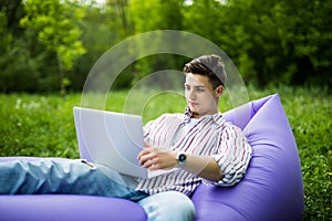 Handsome young man lying on inflatable sofa working on laptop while resting on grass in park
