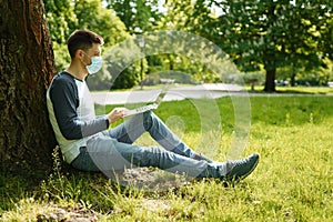Handsome young man in medical mask works on a laptop under a tree in a green park