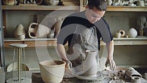 Handsome young man is molding ceramic vase from clay on spinning throwing wheel while working in potter`s studio