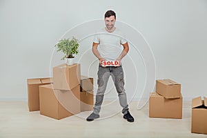 Handsome young man moving into new house, standing, holding plate with red word SOLD written on it, smiling.