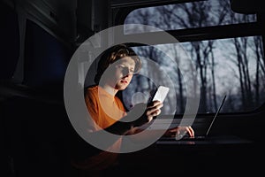 Handsome young man in orange t-shirt uses smartphone on train on the road while traveling, working on laptop