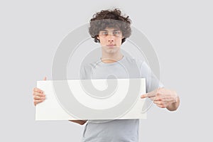 Handsome young man, pointing with finger to show a blank white signboard, isolated on gray background. Placard copy space for text