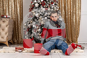 Handsome young man with a scarf sitting with gifts on plaid near the Christmas tree