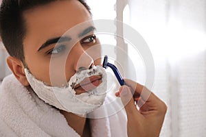 Handsome young man shaving with razor in bathroom, closeup