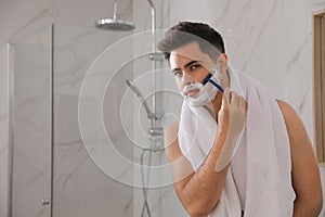 Handsome young man shaving with razor in bathroom, space for text