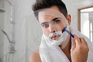 Handsome young man shaving with razor in bathroom, space for text