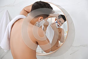 Handsome young man shaving with razor near mirror in bathroom