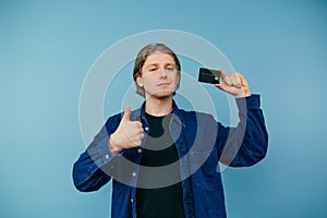 Handsome young man in shirt with bank card in hand, showing thumbs up and smiling looking at camera on blue background