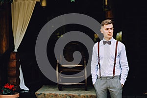 A handsome young man in a shirt, a bow tie, trousers and suspenders stands on the veranda of the house.