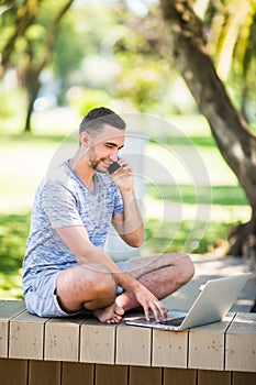 Handsome young man siting on a bench with his laptop on a street next to a park while talking on his phone