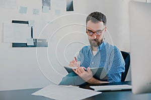 Handsome young man sits in his office while working on business project looks satisfied, takes notes to his agenda, work