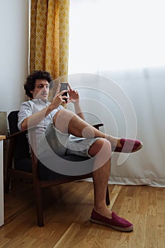 Handsome young man sitting in an armchair with a mobile phone in his hand buying online during quarantine