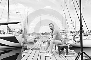 Handsome young man sitting on bench in dock of the bay between boats. Serious boat man on pier looking away