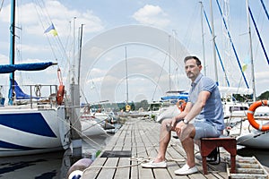 Handsome young man sitting on bench on pier between moored boats. Young businessman on vacation on pier